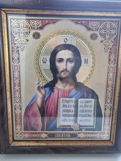The image is a close-up, eye-level photograph of a framed religious icon depicting Jesus Christ. Jesus is shown from the chest up, with a halo around his head. He has dark hair and a beard, and is looking directly at the viewer. His right hand is raised in a gesture of blessing, while his left hand holds open a book. The book has text written in an unfamiliar script. The icon is highly ornate, with decorative borders and symbols in gold and muted colors. The frame of the icon is dark wood with gold accents. The background is a textured, pale gold color. There is no visible indication of the icon's location within the city of Canterbury, United Kingdom.