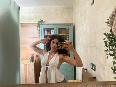 This media file captures a woman taking a mirror selfie in a brightly lit bathroom. She has dark, curly hair and is wearing a white halter top with a plunging neckline, revealing a dark bra and a tattoo on her torso. She is also wearing dark shorts or pants. Her left arm is raised, with her hand touching her head, and her right arm is bent, with her hand holding a smartphone. She is wearing two necklaces, one with a circular pendant and another with a cross pendant, and a ring on her right hand.

The bathroom has tiled walls and a wooden vanity cabinet to the left. A light blue cabinet with glass doors and shelves containing various items, including baskets and bottles, stands against the wall behind her. To the right, a towel is hanging on a rack, and a plant with green leaves is visible. The overall setting suggests a personal, domestic space. The lighting appears to be natural, possibly from a window not directly visible, indicating it could be daytime. The woman's expression is neutral. No visible text is present in the image.