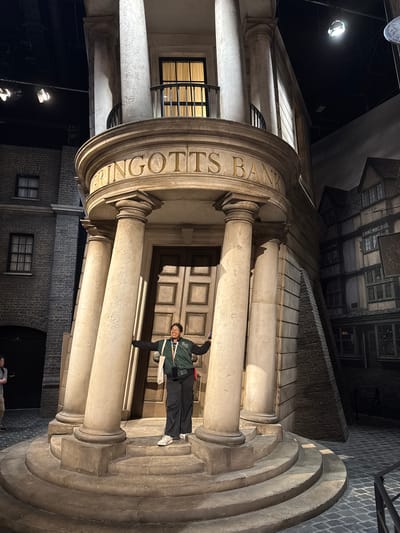 The image shows the facade of Gringotts Bank, a fictional building from the Harry Potter universe, likely a replica or a set in a studio or theme park. The building features classical architectural elements such as large columns, a carved inscription reading "GRINGOTTS BANK," and a massive door with ornate detailing. A person wearing glasses and a green jacket stands in front of the entrance. The surrounding environment suggests a staged or fabricated urban setting, possibly in Nerima, Japan, given the implied location. The light is artificial, highlighting the architecture.