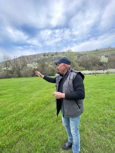 A man stands in a green field, likely in Barkach, Bulgaria, as indicated by the location. The field is vibrant with green grass and interspersed with small white flowers. In the background, a rocky hillside is visible, dotted with trees that appear to be in bloom with white flowers. The sky above is partly cloudy. The man is wearing a blue cap, a black sweater, a gray vest, blue jeans, and dark shoes. He is holding a black object (possibly a bag or folded item) and is extending his other hand towards the field.