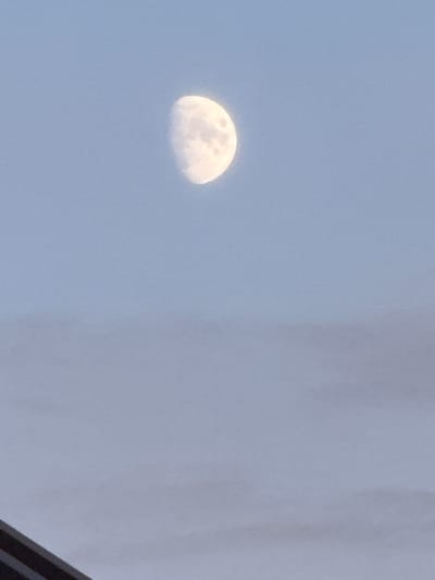 This is a medium-shot, horizontal photograph of the moon in the daytime sky. The moon is positioned in the left half of the frame and appears to be a waxing gibbous phase. It is illuminated and shows some surface detail, appearing bright white with some subtle grey shading, suggesting craters and maria. The sky is a clear, pale blue, indicating it is daytime. To the right of the moon, the sky transitions into a hazy, lighter grey area, possibly due to atmospheric conditions or a very light cloud cover. In the bottom right corner of the image, a dark object with striped patterns, possibly a railing or a building edge, intrudes into the frame, partially obscuring the sky. The overall impression is a clear, bright day with the moon still visible. The time of day is clearly daytime, and the weather appears to be clear or mostly clear with a slight haze. No people or animals are visible. There is no visible text in the image.