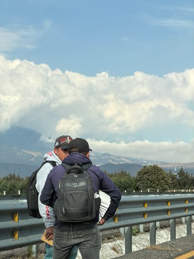Two people stand near a guardrail on the side of a road. One person wears a black hat, backpack, dark shirt, and jeans. The other person wears a baseball cap, gray hoodie, backpack, and jeans. The sky has blue patches and fluffy clouds. Mountains and green trees are visible in the background. The guardrail is metal with yellow reflectors.