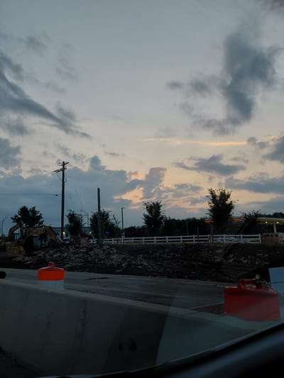 The image depicts a construction scene at dusk in Lexington, United States of America. A large pile of rubble and dirt is visible in the foreground, with heavy machinery, including a yellow excavator, positioned on the left side of the frame. A white picket fence runs across the middle ground, separating the construction area from a road and buildings in the background. Orange traffic cones and a red gas can are present on the road. The sky is filled with dramatic clouds, tinged with the colors of sunset. The overall impression is one of ongoing infrastructure development under a twilight sky.