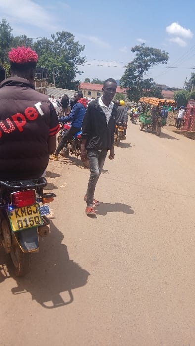 This image depicts a street scene in Kisii Central ward, Kenya, during daylight hours under a bright blue sky with scattered clouds.
In the foreground, the back of a person with distinct bright pink-red dyed hair is visible, seated on a motorcycle. This individual wears a dark, possibly padded, jacket with the red text "Super" horizontally across the upper back. The motorcycle's rear is prominently displayed, featuring a yellow-and-green Kenyan license plate, "KMGJ 015Q." To the right and slightly forward of the motorcycle, a lean young man stands, facing slightly towards the camera with a neutral expression. He is dressed in a dark, possibly black, zipped-up jacket over a light-colored shirt with a dark pattern, dark grey jeans, and red open-toed sandals. Shadows from both individuals and the motorcycle are cast on the light brown, likely unpaved, road surface.
The midground reveals a bustling thoroughfare populated with several other motorcycles and pedestrians. On the right side of the road, a motorcycle is heavily loaded with a substantial stack of long, cylindrical, orange-brown pipes. Further down the street, additional people are visible, some on motorcycles and others on foot. The background consists of dense green tree foliage and several buildings with light-colored walls and reddish-brown roofs, suggesting an urban or semi-urban environment. No specific interactions between individuals are clearly discernible, but the collective presence of people and vehicles indicates a busy public space.
