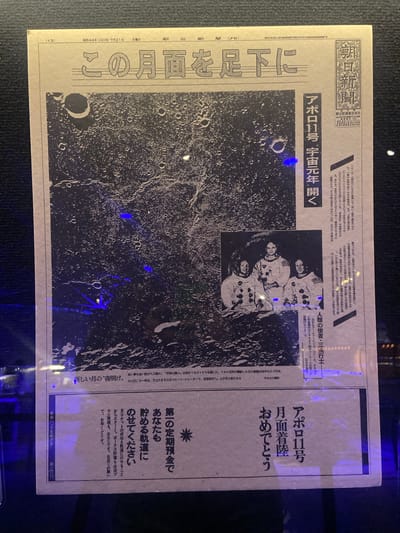 The image shows a framed Japanese newspaper article about the Apollo 11 moon landing. The article features a grainy, black and white photo of the moon's surface with craters, alongside a picture of the three astronauts: Armstrong, Collins, and Aldrin. The newspaper title and other text are in Japanese. The print is encased under glass that has reflections and glare on it, obscuring portions of the print.