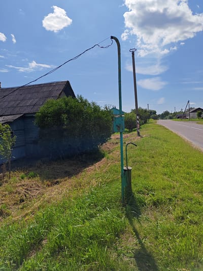 In a rural area near Leninski Rural Council, Belarus, a weathered, teal-colored metal post with a curved top and a hand pump at its base stands in a grassy area. Above the post, a dark electrical wire stretches across the bright blue sky, which is dotted with fluffy white clouds. To the left of the post, a rustic wooden house with a dark, corrugated roof and pale blue siding is partially obscured by lush green foliage. A narrow asphalt road runs along the right side of the image, bordered by a wide expanse of green grass. In the distance, a few more houses and utility poles can be seen, hinting at a small village or hamlet. The scene is bathed in daylight, casting a distinct shadow of the metal post onto the sunlit grass.