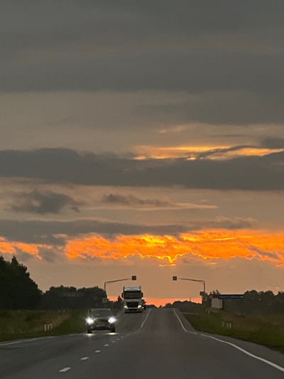 The image depicts a highway scene at dusk with a vibrant orange and yellow sunset visible through dark, dramatic clouds. Two vehicles are on the road: a car with its headlights on in the foreground, and a white truck further up the road, also with headlights illuminated. The road curves uphill, flanked by trees on the left and sparse vegetation and a few structures on the right. Road markers and traffic signals are also present. The location is described as the city of Lermontovskii, Russia.