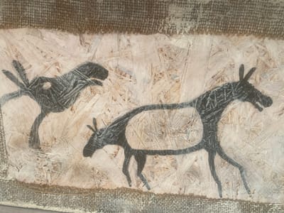 The image depicts cave paintings of two animals in black on a textured, light-colored background reminiscent of rock. The animal on the left is smaller, with outward-pointing appendages. The animal on the right is larger, seemingly equine, with thin legs and a defined outline around its body. The artwork is framed by a rough, burlap-like border along the top and bottom edges. The background appears to be made of pressed wood chips.