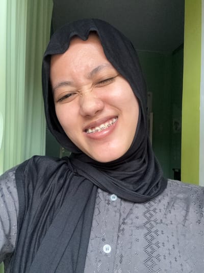 A person wearing a black hijab and a button-up shirt with a textured gray pattern smiles widely, revealing braces. The person's head is tilted, creating a slightly distorted perspective. The background includes a white ledge and a green wall. The overall lighting is soft, and the image has a casual, selfie-like quality. The person's eyes are squinted in a joyful expression.