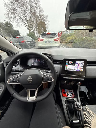 The image depicts a driver's perspective from inside a Renault vehicle, looking out through a rain-splattered windshield at traffic on an overcast day in Mougins, France. The driver's legs, clad in black pants, are visible beneath the steering wheel, with a light-colored slipper on the left foot and a dark shoe on the right. The digital instrument cluster shows a speed of "0," indicating the vehicle is stationary. The infotainment screen on the center console displays a navigation map, identifying the current location as Mougins and the destination as Nice, with the time noted as 14:30. Below the screen are climate controls and a manual 6-speed gear shifter. A red object marked "20" is situated in a console compartment, and a light-colored, patterned blanket or scarf rests on the passenger seat.

Outside the vehicle, a white Nissan crossover with French license plate DJ-450-PN is directly ahead, its brake lights illuminated. To its left, a dark-colored SUV with French license plate CX-072-ZS is visible. The roadside features bare deciduous trees and green bushes, along with a partially visible red and white sign, possibly indicating "Lavage Véhicules." The sky is overcast.