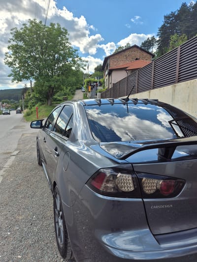 A grey sedan is parked on the side of a rural road in Iskrets, Bulgaria, under a partly cloudy sky. The car, positioned at an angle facing away from the viewer and towards the right, has its rear visible, showcasing tinted windows with reflections of the sky and surrounding trees. A rear spoiler is mounted on the trunk. The road is gravel, and to the right of the car, there is a concrete wall topped with a dark, slatted fence. Behind the fence, a two-story building with a red-tiled roof and brick facade is visible, situated on a hillside covered with trees. In the background, the road winds uphill, with other vehicles and houses visible further down the street. A large tree with green foliage stands prominently on the left side of the road, and power lines extend across the sky.