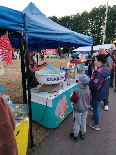 The image shows a street scene, likely a fair or market, in the city of Олтушскі сельскі Савет, Belarus. A vendor, dressed in a striped apron, is operating a cotton candy machine under a blue canopy. The machine is producing blue cotton candy, and the word "СЛАДКАЯ ВАТА" (cotton candy in Russian) is visible on the side of the machine. Several people are gathered around the stall, including a woman in a blue jacket and a child wearing a blue cap, who appear to be customers. In the background, more people, tents, and trees are visible, suggesting an outdoor event. The ground is a paved surface, and the lighting indicates it's likely late afternoon or early evening.