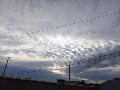 The image captures an overcast sky with streaks of sunlight breaking through the clouds. Power lines crisscross the sky, supported by utility poles. In the foreground, a low, dark wall with concrete pillars is visible. The scene appears to be in the city of Kyzyl Suu, Kyrgyzstan. The overall mood is somber due to the heavy cloud cover, but the sunlight suggests a possibility of clearing weather.
