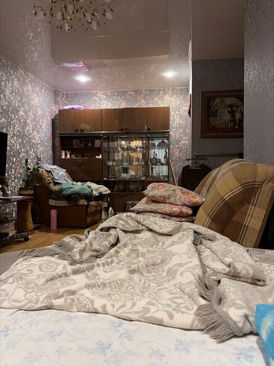 The image depicts a dimly lit living room interior, likely in a residential dwelling in Skopin, Russia. The room is furnished with a large, wooden display cabinet filled with glassware and decorative items, a plush armchair covered with clothing, and several pillows and blankets strewn across a couch or bed in the foreground. The walls are adorned with patterned wallpaper featuring floral motifs, and a large painting hangs on the right wall. A chandelier with crystal-like embellishments is visible on the ceiling, contributing to the overall ambiance. The scene suggests a lived-in space, with items casually placed, indicating everyday use rather than a staged setting. There are no people visible in the image, nor any clear indication of recent activity. The lighting is predominantly artificial and casts shadows throughout the room, adding to the somber atmosphere.