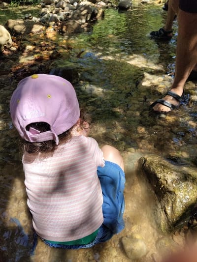 A high-angle, outdoor shot depicts a young child and an adult interacting within a shallow, clear-water stream, likely within the Maale Yosef Regional Council, Israel. The child, viewed from behind, is seated directly in the water, wearing a light purple baseball cap with yellow accents, a light pink and white horizontally striped short-sleeved top, blue shorts, and a visible green undergarment. The lower legs and feet of an adult, wearing dark open-toed sandals, are visible standing in the water to the right of the child. The stream bed consists of numerous smooth, light-colored stones and pebbles, clearly visible through the transparent water. Dappled sunlight filters through an unseen canopy above, creating contrasting light and shadow patterns on the water's surface and the submerged rocks. A larger, dark, possibly moss-covered rock is partially submerged to the child's right. The scene indicates an interaction or activity taking place within a natural water environment.