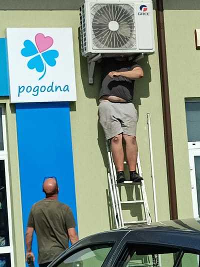 In this photograph, a man is seen working on an air conditioning unit mounted on the side of a building. He is standing on a ladder, with his upper body obscured by the air conditioner itself. He is wearing a dark t-shirt and light-colored shorts, with black shoes. Another man, wearing a dark olive t-shirt and sunglasses on his head, stands below and to the left, looking up at the air conditioner.

The building has a light green facade. To the left of the air conditioner is a sign with a white background, featuring a blue four-leaf clover with a pink heart in the center. Below the clover, the word "pogodna" is written in blue cursive. Beneath the sign is a vertical blue panel. A dark-colored car is partially visible in the foreground, with its roof and hood taking up the bottom portion of the frame.

The scene appears to be outdoors on a sunny day, indicated by the strong shadows cast by the ladder and the man working. The setting seems to be a commercial or residential area in Czyżew, Poland. The activity suggests installation or maintenance of the air conditioning unit. The overall mood is one of a typical daytime scene, with a focus on the task at hand.