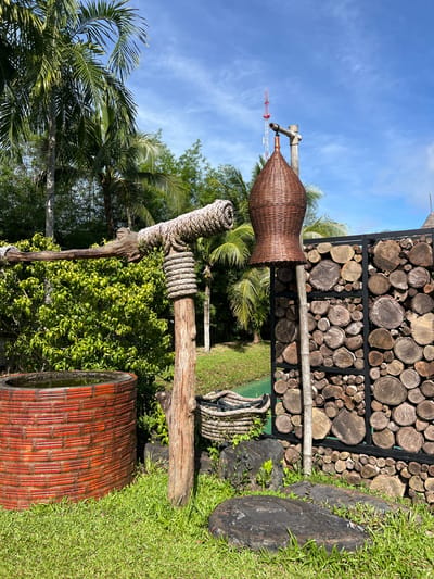 The media file displays an outdoor scene in a tropical setting, likely Thailand, given the presence of lush palm trees and dense green foliage. The image is oriented vertically, giving a sense of looking up.
In the foreground, there are several rustic, naturalistic decorative elements. A large, woven basket, shaped like a wide-mouthed bottle, is supported by wooden posts wrapped with thick rope. Another similar rope-wrapped wooden post is visible below it. To the right, a large, round, terracotta-colored planter with a woven pattern sits on vibrant green grass. The planter appears to be empty or filled with something reflective that mirrors the sky.
In the background, a wall constructed from stacked, circular wooden logs of varying sizes is a prominent feature. This wall is framed by dark metal supports. Beyond the wall, more tropical vegetation and palm fronds are visible, suggesting a garden or natural park setting. A tall, red and white lattice tower, possibly a communication tower, is visible in the distance against a bright blue sky with scattered white clouds.
The time of day appears to be daytime, with clear blue skies and bright sunlight casting shadows, indicating fair weather. There is no text visible in the image. The overall impression is one of a serene, natural, and possibly tropical garden or outdoor recreational area.