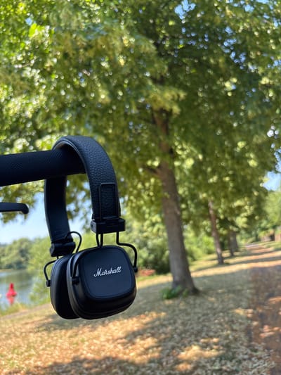 A pair of black Marshall headphones are hanging from a black textured bar, with the "Marshall" logo visible on the right earcup. The background is a sun-dappled park with lush green trees and a body of water on the left. The ground is covered with dry leaves and grass. The scene appears to be an outdoor recreational area. The location is Erlenbach am Main, Germany.