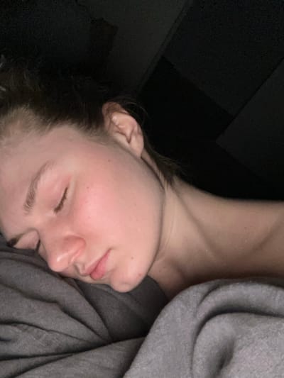 The image presents a close-up view of an individual, identified as a young adult female, in a state of sleep or deep rest, within a location specified as Бакча-Сарай, Russia. The subject is oriented on her left side, with her left cheek and the side of her face resting on a grey fabric, consistent with a pillow or bedding. Her eyes are closed, and her facial features appear relaxed. Dark brown hair is visible, pulled back from the forehead and around the ear. The skin is fair-toned, with a slight pink flush visible on the left cheek. The bare neck and upper right shoulder are partially visible, exhibiting a subtle sheen or glistening, possibly indicative of perspiration or light reflection. A grey blanket or bedding covers the lower portion of the subject and extends to the lower left of the frame. The background is uniformly dark and devoid of any discernible objects or architectural features, suggesting a dimly lit indoor environment. No other individuals or interactive elements are present in the scene.