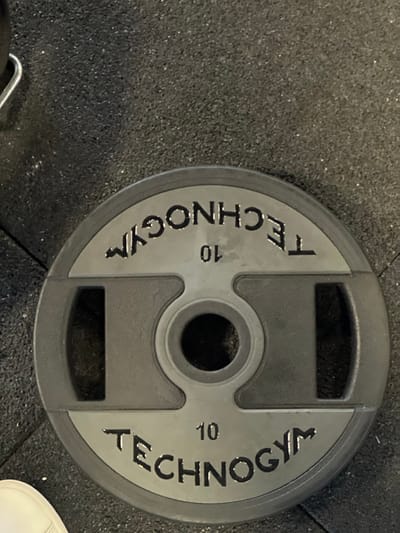 A dark gray circular Technogym brand weight plate, marked with "10" (presumably kilograms) and "TECHNOGYM" text embossed twice on opposing sides, rests on a dark, granular rubber gym floor in Wolfisheim, France. The weight plate features three handle cutouts, with a central bore for a barbell. A white shoe toe is visible in the lower-left corner of the frame, and a silver metal hook or handle of an unidentified object is partially visible in the upper-left. No individuals are fully depicted, and no explicit actions or interactions are occurring.