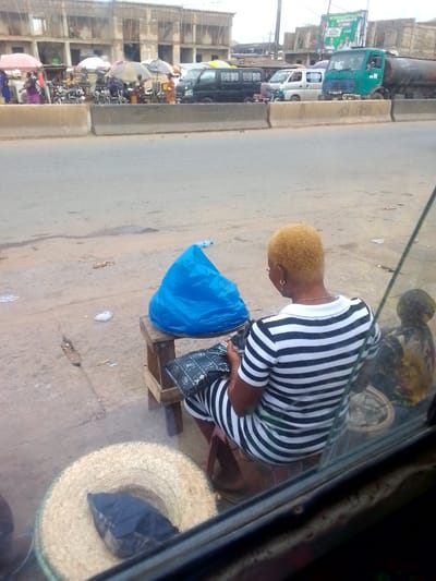The image depicts a street scene in Ihiala, Nigeria. A woman with short, bleached hair is seated on a small wooden stool, wearing a black and white striped dress. She is holding a black patterned bag in her hands. Next to her is a blue plastic bag placed on another small stool. In front of her, partially obscured by the camera's vantage point through a vehicle window, is a straw hat.
Across the road, behind a concrete barrier, is a bustling market scene. Several people are visible along with motorcycles and parked vans. A green truck is prominent in the background. Buildings under construction are also visible in the distance. The weather appears overcast.