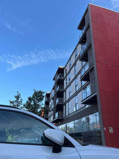 The image shows a modern apartment building with a bright red section on the right side, in Milton Keynes, United Kingdom. The building has multiple floors with balconies and large windows. A white car is parked in front of the building, partially visible in the foreground. The sky above is blue with wispy white clouds. Trees are visible behind the car and to the left of the building. A security camera and a smoking ban sign are mounted on the red wall of the building. The overall impression is a contemporary urban scene.