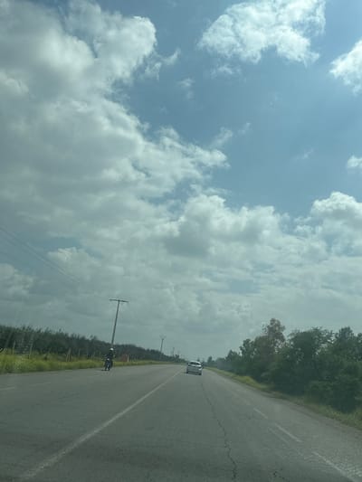 A slightly overexposed photograph shows a country road near Saâda, Morocco. The road stretches into the distance, a white car visible far ahead. A motorcycle with a rider is visible on the right side of the road, traveling in the same direction as the car. Utility poles with wires run along the right side of the road. The left side of the photograph is dominated by a sky filled with white, cumulus clouds. Trees and shrubs line the road on both sides. The asphalt shows a white dividing line.