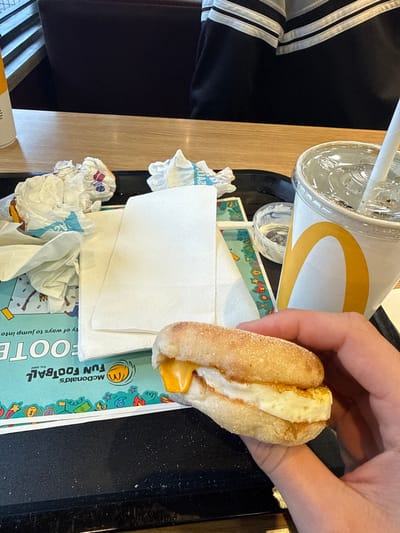 The scene depicts the interior of a McDonald's restaurant in East Hampshire, United Kingdom. The primary focus is a black plastic serving tray situated on a light-colored wooden table. A human hand, positioned in the lower right, holds a breakfast sandwich composed of an English muffin, a folded fried egg patty, and a slice of melted orange cheese.
On the tray, a McDonald's branded paper cup with a clear plastic lid and a white straw contains a beverage. A stack of clean, white paper napkins rests beside several crumpled paper wrappers, some exhibiting blue text, indicating partially consumed items. A small, clear plastic ramekin containing a white substance and a small spoon is also visible. The tray liner beneath these items is a blue and white paper placemat, partially visible and oriented inverted, displaying "McDonald's Fun Football est. 2002" branding.
In the background, a person is partially visible from the mid-torso down, wearing a black garment with two horizontal white stripes on the sleeve, seated in a dark brown upholstered booth or chair at the same wooden table. The environment is typical of a fast-food dining area.