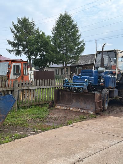 The image depicts an outdoor scene with agricultural machinery and residential buildings. In the foreground, a blue tractor with a front-mounted plow is parked on a muddy patch of ground, adjacent to a concrete path. To the left, an orange tractor is partially visible behind a weathered wooden fence. Several trees, some appearing to be coniferous, are situated in the background. Behind the tractors and fence, there are houses with dark roofs, suggesting a rural or semi-rural setting. Overhead, power lines crisscross the sky. The overall environment appears to be somewhat overgrown with grass and includes a small puddle of water. A dark-colored animal, possibly a cat, is visible near the fence. The image is taken from a ground-level perspective, looking slightly upwards towards the machinery and buildings. The sky is overcast with clouds. This scene is consistent with a location in the city of Чайковский, Russia.
