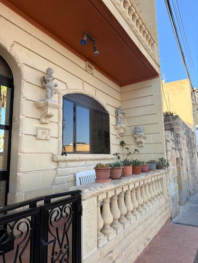 The image depicts the exterior of a building on a sunny day in Xagħra, Malta. The building has a light-colored stone facade with decorative elements. A balustrade runs along the side of the building, with potted plants placed on top. There are two statues of cherubs mounted on the facade, one above a carved house number "51" and the word "THEUMA". A dark-colored metal gate with an ornate design is visible in the foreground. The background shows a clear blue sky and adjacent buildings. The scene is static and appears to be a residential or commercial property.
