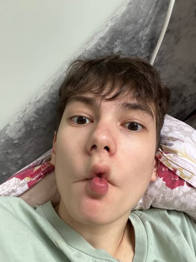 An individual is depicted in a close-up, front-facing selfie, taken indoors, presumably in Zainsk, Russia. The subject, with short brown hair and light skin, gazes directly into the camera with wide-set brown eyes. Their lips are intentionally pursed and pushed outwards, forming a circular protrusion. The individual wears a light green crew-neck garment, with a thin silver chain visible at the base of the neck. The background consists of a grey, textured, possibly upholstered surface on the left and a pillow with a patterned pillowcase, featuring red and maroon floral elements on a light ground, on the right. A portion of a light-colored wall is visible in the upper left.