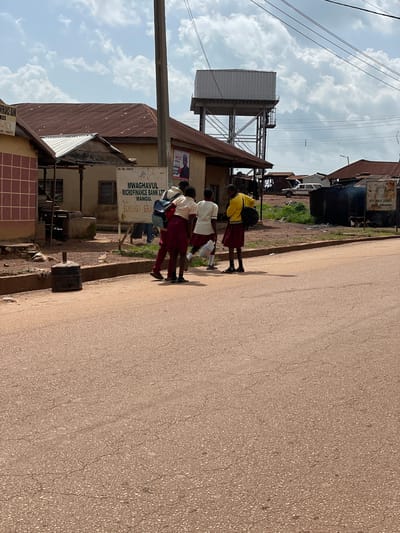 The image captures a street scene in Tangu, Nigeria, under a partly cloudy sky. In the foreground, a paved road with a reddish-brown surface exhibits minor cracks. Along the left side of the road, a concrete curb separates the pavement from a dusty area with sparse vegetation. To the left, a building with a tiled facade and a corrugated iron roof is visible. A sign on this building reads "FINANCE BANK, KOS BRANCH, NO. 196673."

In the middle ground, a group of four young people, likely students, are standing near a sign that announces "MWAGHAVUL MICROFINANCE BANK LTD MANGU, BOKKOS BRANCH, RC. NO. 196673." Three of the individuals are wearing school uniforms consisting of white shirts and maroon skirts or trousers. One student in a yellow top and maroon skirt carries a backpack. The students appear to be interacting or waiting.

Behind the microfinance bank sign, a modest building with a corrugated iron roof and beige walls is present. Further back, a large elevated water tank with a metal platform stands against the sky. The background reveals more buildings with corrugated iron roofs, some appearing weathered, and the presence of power lines overhead. A white car is parked in the distance. The overall environment suggests a somewhat rural or developing urban setting.