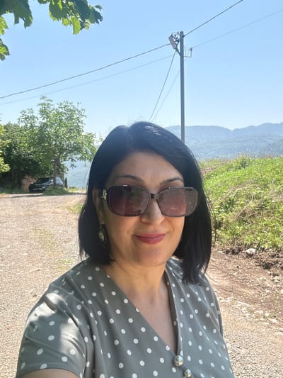An outdoor, close-up, waist-up, selfie-style portrait shows a woman with dark hair and sunglasses against a backdrop of green hills and trees under a clear blue sky. She is wearing a grey, polka-dot dress with silver buttons. The ground is a gravel path. In the background, a black SUV is parked near a building partially obscured by trees. A utility pole with wires is visible in the mid-ground. The scene is likely located in or near Yenokavan, Armenia, known for its scenic mountainous terrain.