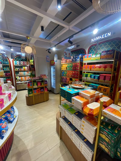 The image depicts the interior of a brightly lit sweet and namkeen shop. The shop is well-stocked with a variety of packaged snacks and sweets displayed on shelves and in boxes. Prominent signage indicates sections for "BAKERY" and "NAMKEEN." Several products feature colorful packaging, with text in English and Hindi visible on some. A prominent refrigerator unit is visible in the background. The flooring appears to be light-colored wood or laminate. The overall environment suggests a commercial establishment focused on food retail.