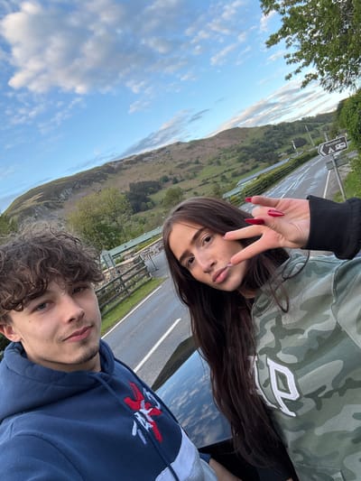 Two individuals are visible in the foreground of the photograph. One person, in the lower portion of the image, has curly brown hair, fair skin, and is wearing a blue hoodie with a red and white design. The other person is positioned above, wearing a camouflage pattern hoodie with a white logo, black sleeves and red painted nails. A peace sign gesture is performed. Behind them is a paved road with white lane markings and a green roadside barrier. Further back, a large, grassy hill or mountain is visible against a blue sky with scattered white clouds. A sign can be seen near the top of the frame with 'Henstent' on it. This road is located near Llangynog, United Kingdom.