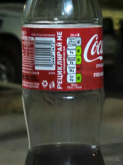 A close-up, low-angle shot shows the label of a clear plastic bottle of Coca-Cola. The label is red with white text and graphics. The Coca-Cola logo is prominently displayed on the right side of the label, in white cursive font. To the left of the logo, there is a nutritional information panel with values for energy, fat, and sugar, presented with green and white icons. A barcode is also visible on the left side of the label. Below the nutritional information, there are recycling symbols and a trash bin icon. The bottle appears to be partially filled with a dark liquid. The background is out of focus and appears to be an indoor or outdoor setting with muted colors. There is no specific indication of the city of Biaga, Bulgaria in the image.