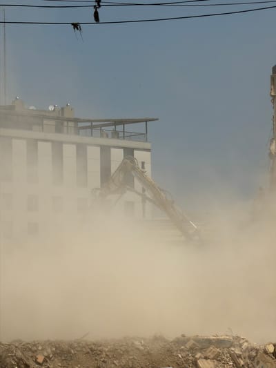 An excavator is seen demolishing a building in a cloud of dust. The building appears to be a multi-story structure with a white facade and a rooftop balcony. Several satellite dishes are visible on the roof. In the foreground, a pile of rubble and debris is present. Overhead power lines are strung across the top of the image. The sky is a clear blue.