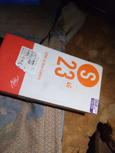 The image shows a box for an itel S23 4G smartphone. The box is primarily white with red on the left side where the brand name "itel" is displayed. Prominently featured is the text "S 23 4G" in orange, followed by "50MP AI Rear Camera". A purple sticker on the right corner indicates "8GB RAM (4+4GB)" and "128GB ROM". A white label on the left displays manufacturing details. The box is placed on a patterned cloth surface.