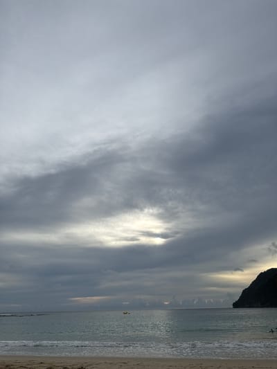 This is a wide-angle, eye-level shot of a beach scene on a cloudy day, likely in Meunasahbale, Indonesia. The sky is filled with heavy, gray clouds, with a bright patch of sunlight breaking through in the middle distance. The sea is calm with gentle waves lapping at the sandy shore. In the water, a small yellow boat is visible in the distance. To the right, a dark, tree-covered hill juts out into the sea. There are two people visible in the water near the shore on the right side of the frame, with one appearing to be a swimmer. The sand in the foreground is a light brown color, with some subtle ripples from the receding tide.