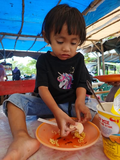 In Meurah, Indonesia, a young child, likely a girl with dark, straight bangs, is seated on a plastic-covered surface, engaged with food at what appears to be an informal outdoor or semi-outdoor eating establishment. The child wears a black short-sleeved t-shirt featuring a colorful unicorn graphic and light blue denim jeans. Her right bare foot, showing minor dirt marks, is visible.
The child is focused on an orange plastic plate containing instant noodles (identifiable by their appearance and the "Pop Mie Kari Ayam" cup nearby) and is holding a light-colored, irregularly shaped food item, possibly a cracker or a piece of fried food, over the noodles. A white plastic utensil rests on the plate. A stack of two yellow "Pop Mie Kari Ayam" instant noodle cups is positioned to the right of the child. An adult's arm, holding a similar orange plate, is partially visible to the child's right.
The overhead structure consists of a blue tarp covering a wooden or bamboo frame, with sections of a white/beige tarp visible behind it, suggesting a temporary shelter. In the background, a red barrier or counter separates the immediate eating area from a food preparation or service area. Two blurry figures of adults, one in a pink headscarf and another in a dark headscarf, are visible near a display stand with various items. Further in the background, green plastic chairs and a green school bus ("BUS SEKOLAH") are discernible, indicating proximity to a road or public space. The child sits on a surface covered by a translucent plastic sheet, revealing a subtle pattern beneath.