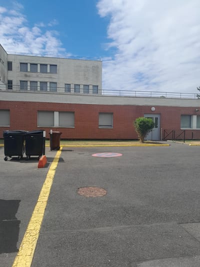 This is a full-shot, eye-level, outdoor photograph taken on a cloudy day in Roissy-en-Brie, France. The scene depicts the exterior of a building with a brick lower section and a light-colored, ribbed metal upper section. A yellow line is painted on the asphalt in the foreground, extending diagonally from the lower left to the center of the image. To the left of the yellow line are two large, black wheeled garbage bins. A smaller, brown wheeled bin is situated to the right of the black bins. An orange traffic cone rests near the yellow line.

The brick facade of the building features several windows with grey, slatted blinds. A single, circular outdoor light fixture is mounted on the brick wall above a grey door, which is partially obscured by a dense, green bush. A short, black metal fence runs along the sidewalk in front of the building. A circular red and blue sign with a white symbol is painted on the asphalt near the bush. Further back, the upper portion of the building is visible, characterized by multiple windows with grey frames. The sky above is blue with scattered white clouds. A railing is present along the top of the building. The image captures a typical urban environment, likely a commercial or institutional setting.
