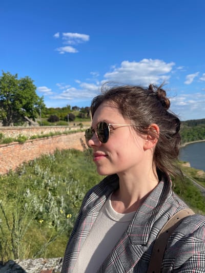 This image presents a close-up, side-profile portrait of a young woman enjoying an outdoor setting on a sunny day, taken from an elevated perspective. The woman, with her dark hair styled in a messy bun, is wearing round, gold-rimmed sunglasses. Her head is tilted slightly downwards and to the right, showing her left profile from her chin to her ear. She has fair skin and a visible dimple on her cheek. She is dressed in a white tank top, layered under a grey and black plaid blazer featuring subtle red accents. A light brown belt is visible over her shoulder, suggesting it's part of her outfit or a bag strap.
The background unfolds into a bright blue sky with scattered white, fluffy clouds on the left. Below, a lush green landscape includes an old, weathered brick wall or structure running diagonally across the mid-ground, partially covered by vegetation. Beyond this, a wide expanse of green grass dotted with small yellow wildflowers leads to a body of water, likely a river, visible in the distant background. Distant green hills and trees line the far bank of the river. The scene suggests a historical site or fortress overlooking a river, aligning with the provided location context of Petrovaradin, Serbia. The lighting indicates bright daylight. The woman appears relaxed, gazing downwards at the scenery. No text is visible in the image.