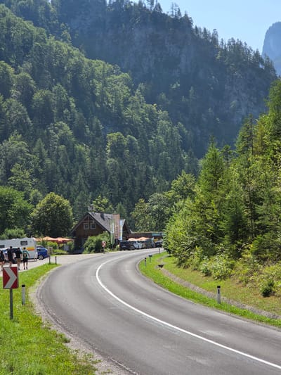 The image captures a scenic view of a winding asphalt road in Landl, Austria, flanked by lush green trees and a towering, rocky mountain face. On the left side of the road, a building with a brown wooden exterior and a dark roof is visible, featuring several windows and a satellite dish. In front of the building, outdoor seating with orange umbrellas is set up. A few cars, a caravan, and a small group of people are parked or standing near the building. The road itself is marked with a solid white line down the center and is bordered by grassy verges and reflective posts. The overall scene suggests a tranquil, natural environment, possibly a stop along a scenic route.