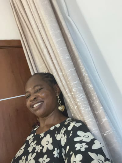 A woman with braided hair and gold heart-shaped earrings smiles at the camera. She wears a black dress with white flower patterns. Behind her, there are cream-colored curtains and a brown wooden door. There is also a white electrical cord hanging in front of her face. I cannot determine the location in Mopo-Akinlade, Nigeria.