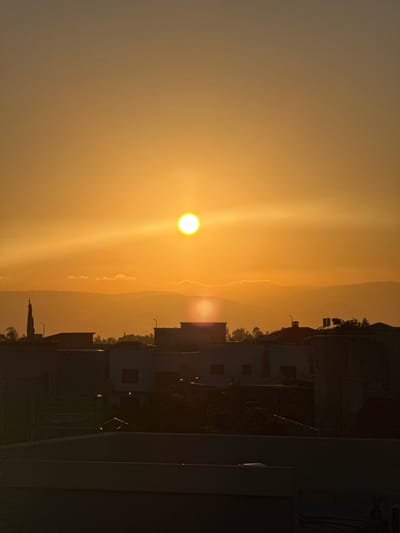 This media file is a vertical, full-frame photograph taken at sunset in Katzrin, Israel. The scene captures the sun as a bright, radiant orb positioned in the left half of the frame, casting a warm, golden glow across the sky. Streaks of light emanate from the sun, illuminating thin layers of clouds and the distant silhouette of mountains.
In the right half of the image, the dark silhouettes of buildings and trees are visible. These structures appear to be residential, with flat roofs and some visible windows. The architecture suggests a Middle Eastern setting. A narrow, spire-like structure, possibly a minaret of a mosque, is discernible amongst the buildings. The overall impression is one of a peaceful evening in a town nestled against a natural landscape. The image evokes a sense of tranquility and the beauty of a natural phenomenon. No people are visible in the frame, and there is no visible text. The weather appears clear, with the sunset being the dominant atmospheric feature.