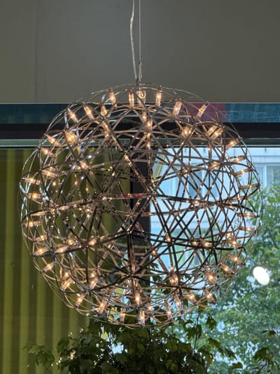 A spherical, metallic chandelier with multiple lights is suspended from the ceiling in a room with a window. The chandelier is made of interconnected metallic strips forming a geodesic dome-like structure, with small lights embedded along the strips. Through the window, a blurred view of greenery and a building is visible. There are no people or specific actions depicted in the image. The location in the city of Yukki, Russia is not discernible from the image itself.