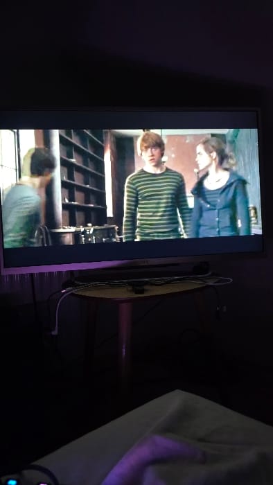 The image depicts an indoor scene within a residence in Heusden, Belgium, observed from a low angle. The room is dimly lit with a prominent purple ambient light source originating from the left. A flat-screen television, identified by a "SONY" logo on its lower bezel, dominates the upper half of the frame.

The television screen displays a scene from a film or television show featuring three individuals. In the center, a male character with reddish-brown hair wears a green and dark-striped long-sleeved shirt, facing forward with an open-mouthed expression of surprise or reaction. To his right, a female character with brown hair tied back, dressed in a dark blue hooded jacket over a striped shirt, looks towards the central male. On the left, a third male character, seen from the side or slightly from behind, wearing a light-colored long-sleeved shirt, is partially visible. The background of the on-screen scene features tall, dark wooden shelving units, indicative of a library or study.

Below the television, a light-colored, possibly pinkish-brown, rectangular table with tapered legs is positioned. Multiple black and white electrical cables are coiled and draped across the table's surface. In the bottom-left foreground, a portion of a human hand is visible, illuminated by the purple light, resting on a light-colored, soft surface, likely bedding or a blanket. Adjacent to the hand, a small electronic device, possibly a game controller, shows a faint blue glowing light and another pink glowing light.