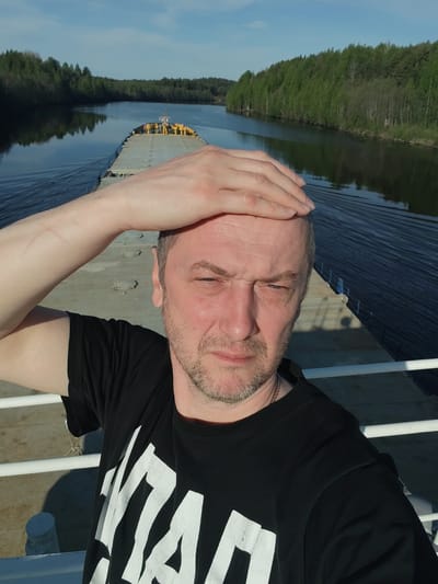 A medium, eye-level selfie of a middle-aged man on a barge moving through a canal. The man has light skin, short grey hair, and a grey beard. He is wearing a black t-shirt with a white design. His right hand is on his forehead, shielding his eyes. The barge is grey with a yellow structure at the far end. The canal is lined with green trees reflecting in the water. The sky is blue with some white clouds. This could be the Volga-Baltic Waterway, given the reference to Depo, Russia.