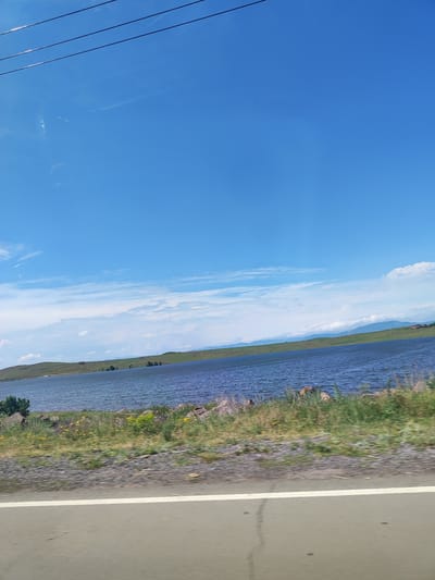 The image depicts a wide, calm body of water, likely a lake, under a bright blue sky with scattered white clouds. A green, rolling hillside forms the distant shore, dotted with sparse trees and some structures, possibly houses or outbuildings, in reddish hues. The foreground features a grassy embankment with some rocks and small yellow flowers, transitioning to a paved road with a white line marking its edge. Overhead, three dark power lines stretch across the top of the frame. The overall scene suggests an outdoor, natural setting, possibly a reservoir or a large lake near a semi-rural or hilly area. The location is not specified as Horom, Armenia within the image itself.