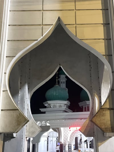 The image shows a view of a mosque in Siron, Indonesia, framed by a decorative architectural element. The foreground features a curved, pale archway with textured surfaces and visible rivets. Beyond the archway, the mosque's features are visible against a dark night sky. These include a prominent green dome, topped with a spire, and a white minaret with arched windows. Further in the background, more white architectural elements and arches of the mosque complex are illuminated. Red lettering is visible on a sign within the mosque's structure. Several people are present in the background, appearing to be walking or standing within the mosque grounds. The overall lighting suggests it is nighttime.